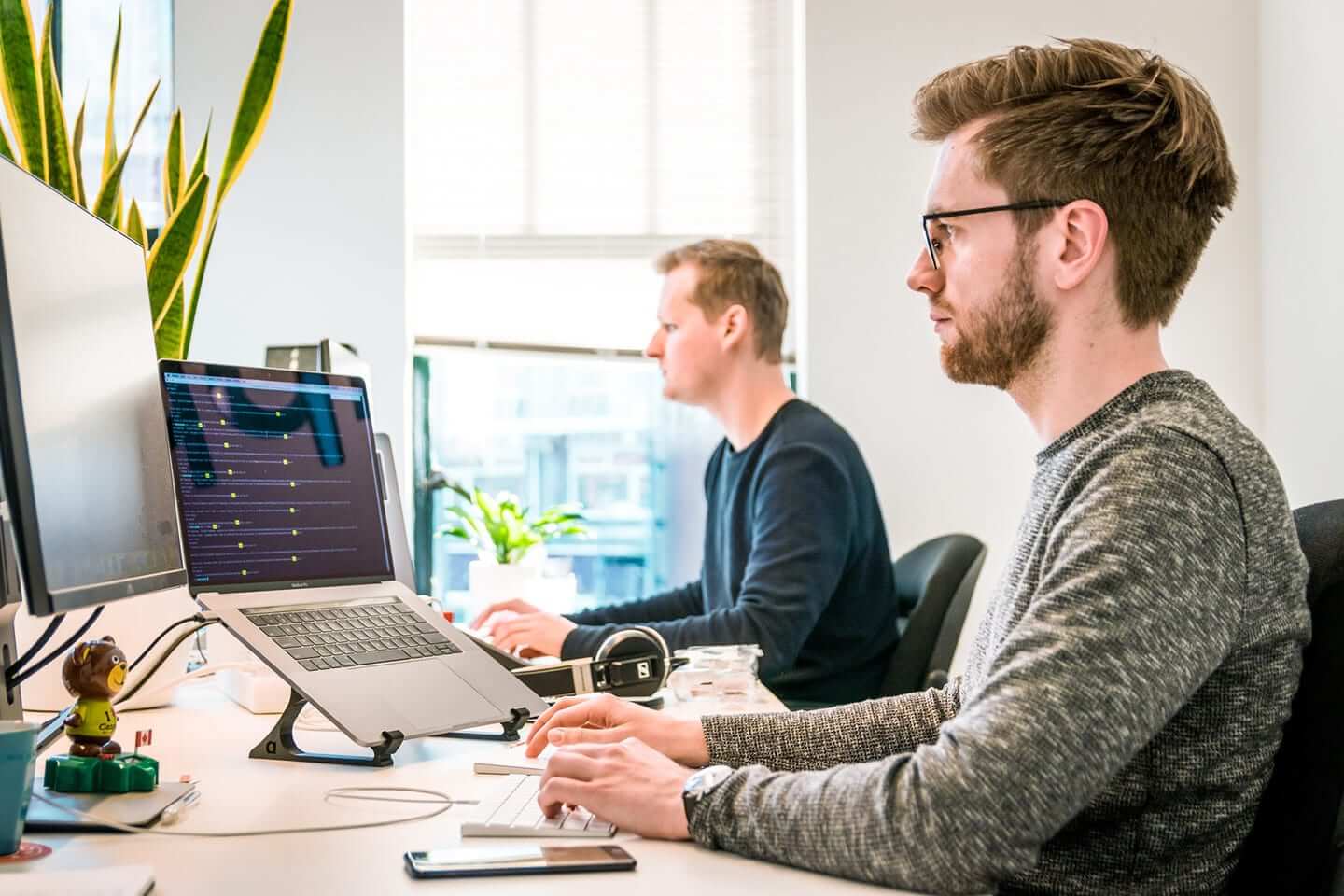 Two people working at laptops in a bright office, one typing with code on screen